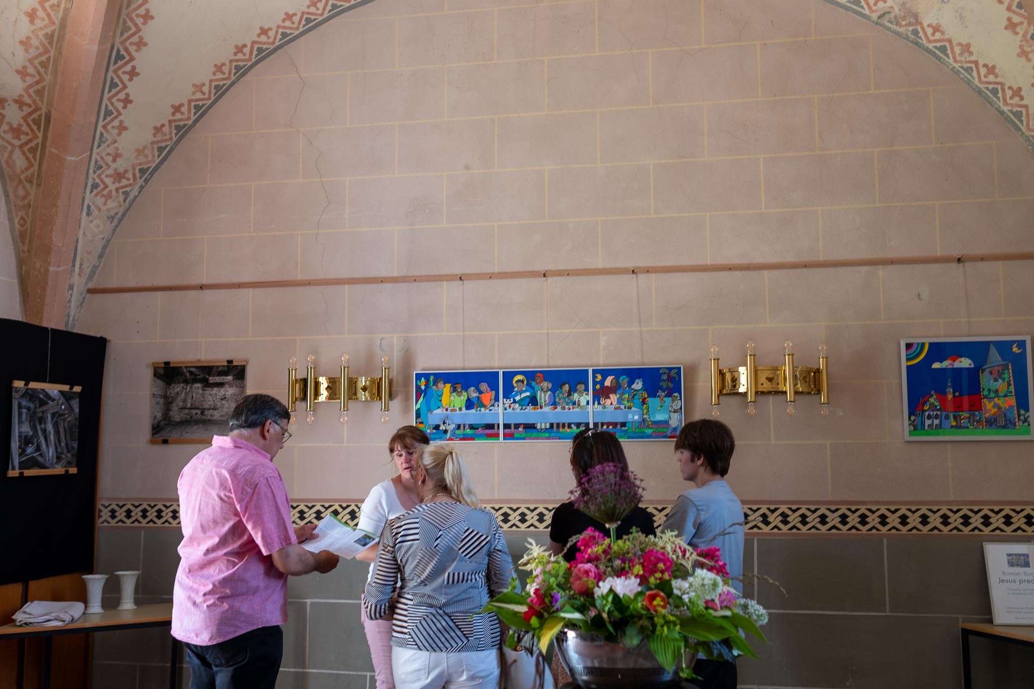 5 Personen stehen im Kreis im Innenraum einer Kirche. Eine männliche Person schaut dabei in ein aufgeschlagenes Buch. Im Vordergrund steht ein Blumenstrauß. im Hintergrund befinden sich einzelne Bilder und Leuchten.