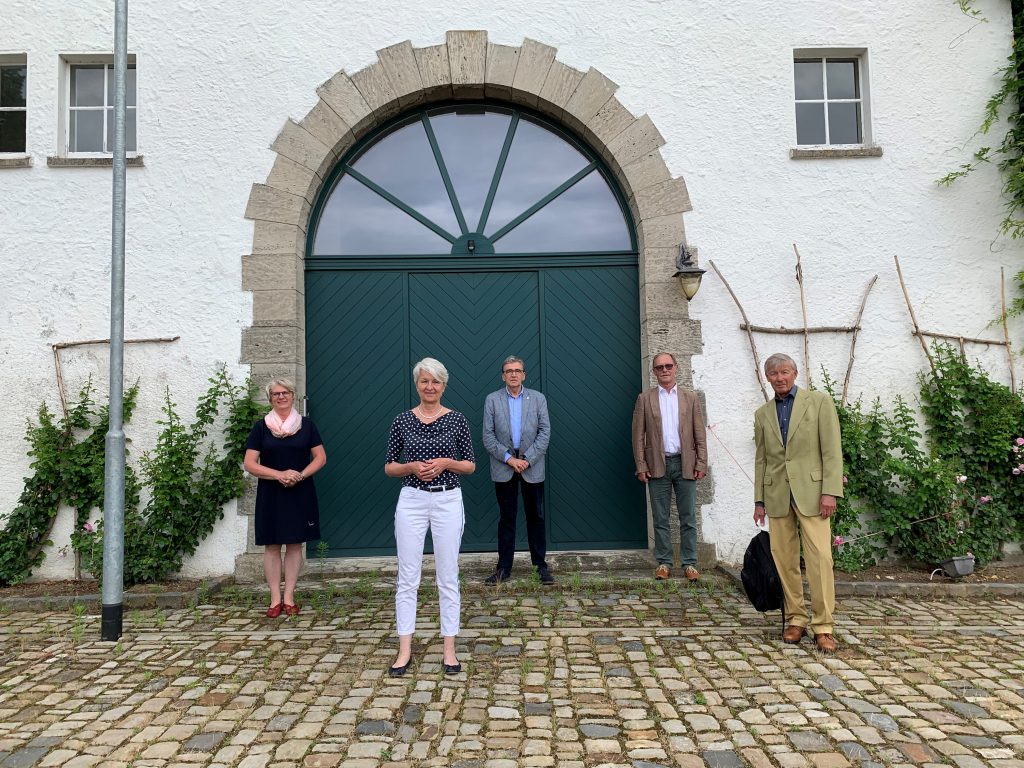 Fünf Personen stehen vor einem weißen Gebäude, vor einer grünen Eingangstür als Rundbogen.
